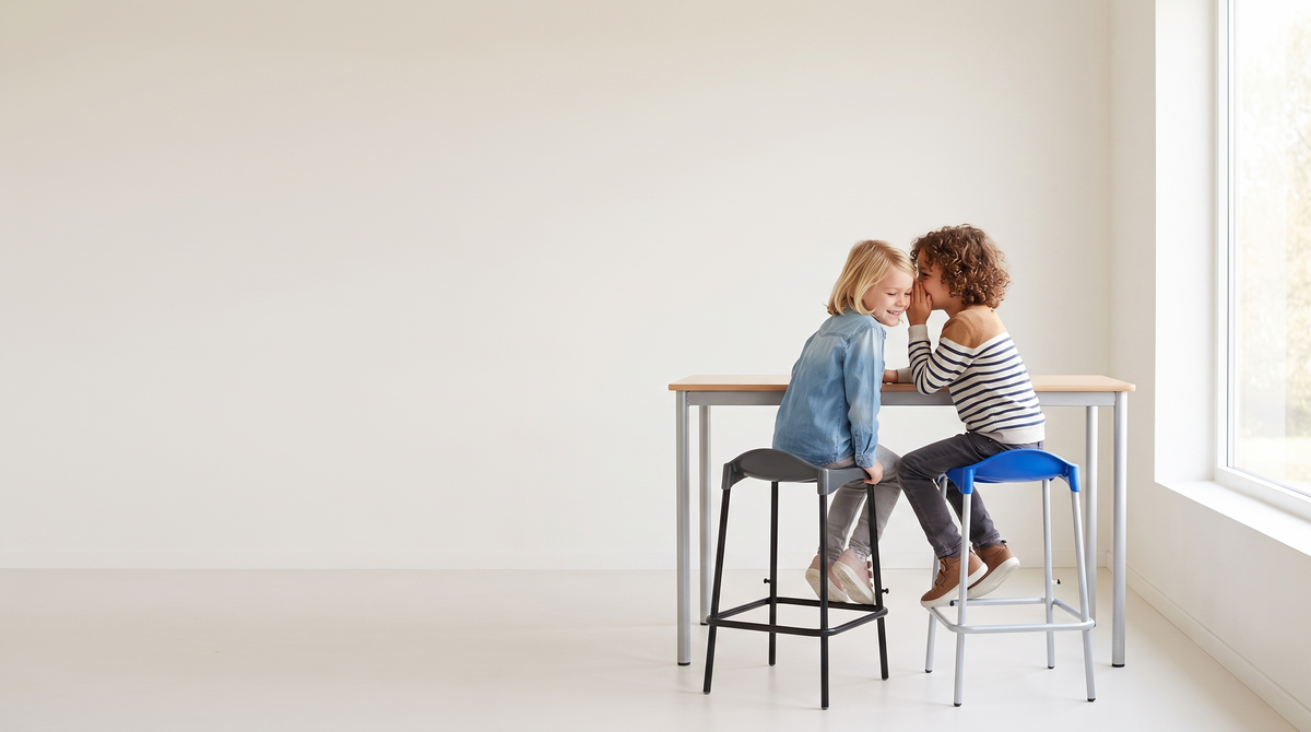 Two children sitting at a small table on stools in a bright, minimal room with a large window and off-white wall.